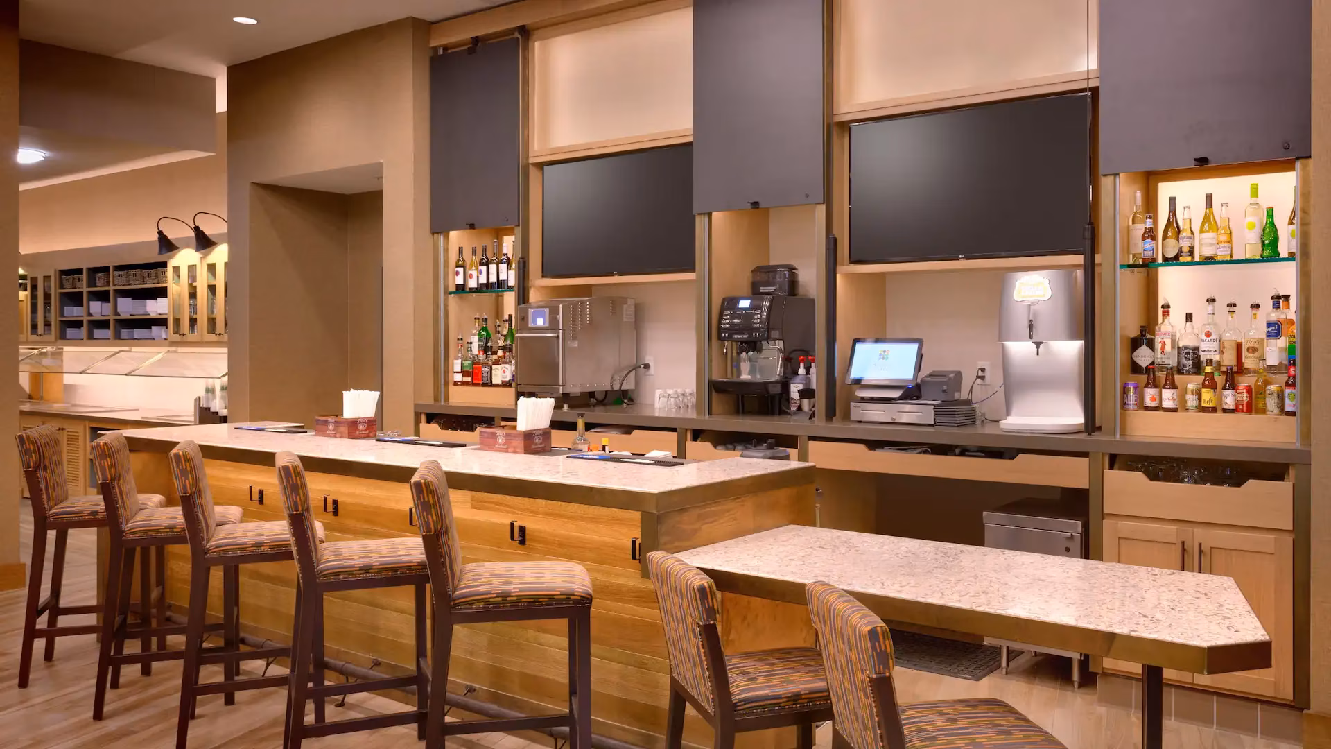 Modern bar area with stools, televisions, drinks, and coffee machines.