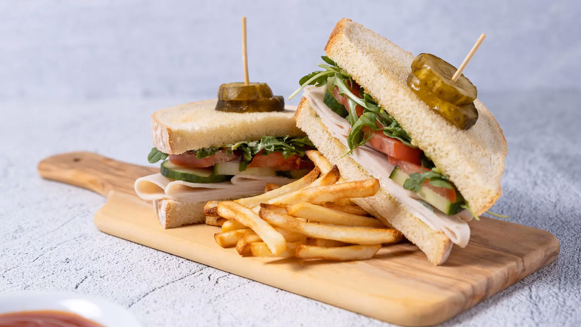 Two deli meat sandwiches, fries, and pickles on a cutting board.