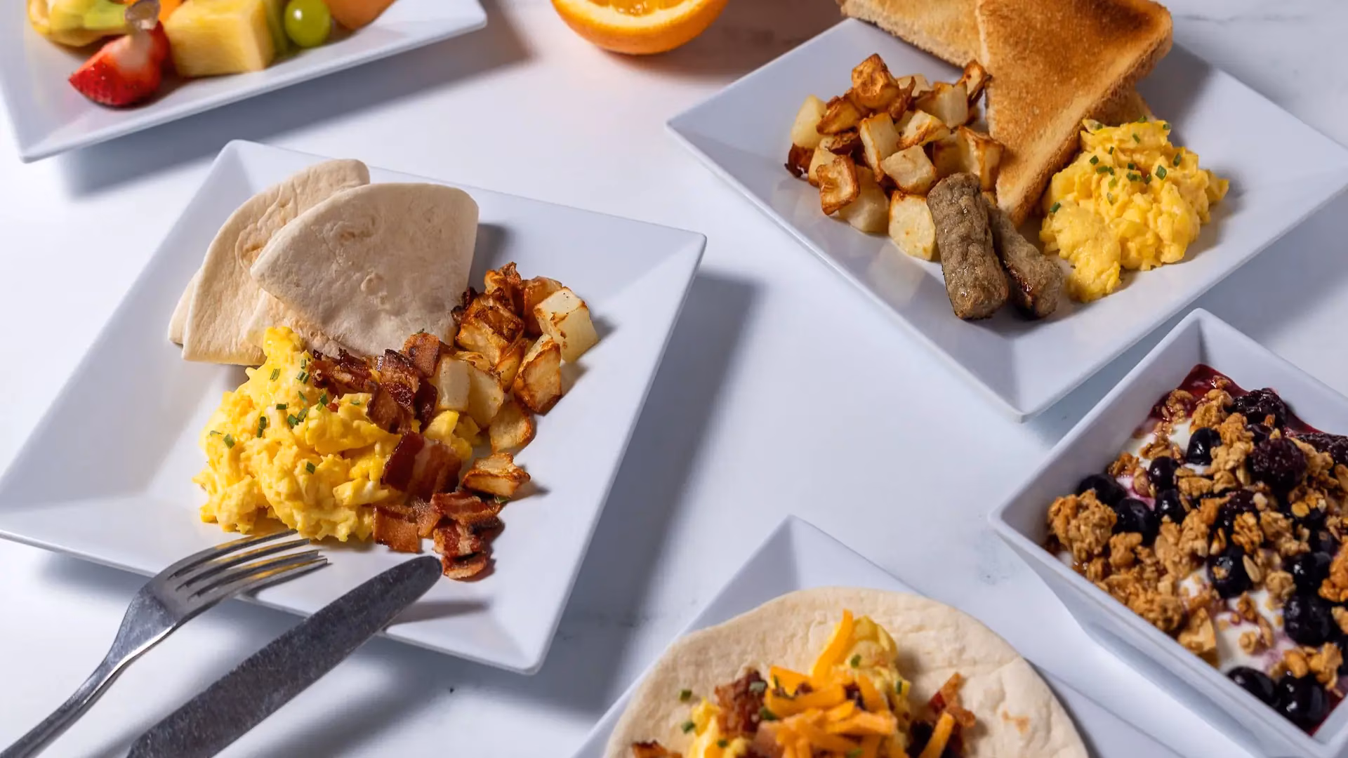 Breakfast plates with scrambled eggs, potatoes, meat, fruit, and yogurt.
