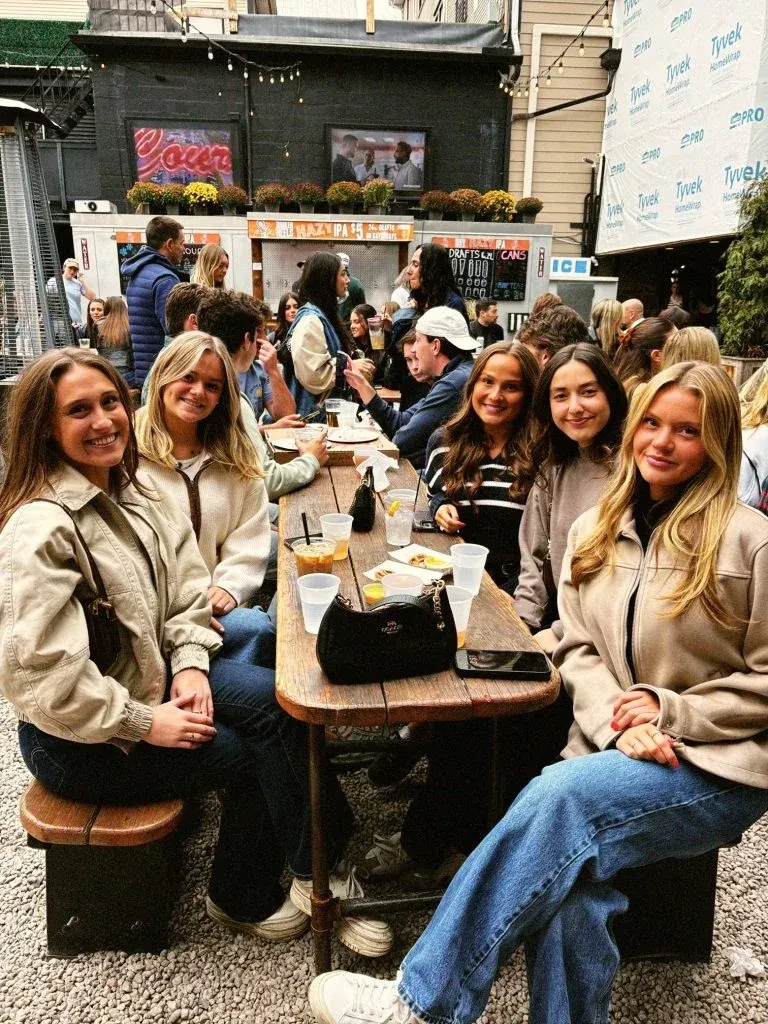 Four smiling women with drinks at a busy outdoor beer garden.