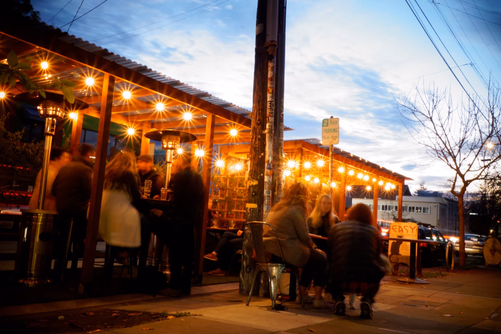 Outdoor patio with people, glowing string lights, and heaters at dusk.