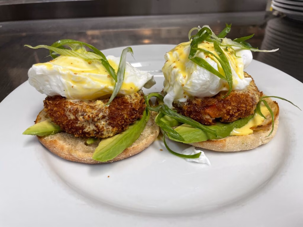Two English muffins with breaded patties, avocado, poached eggs, sauce, scallions.