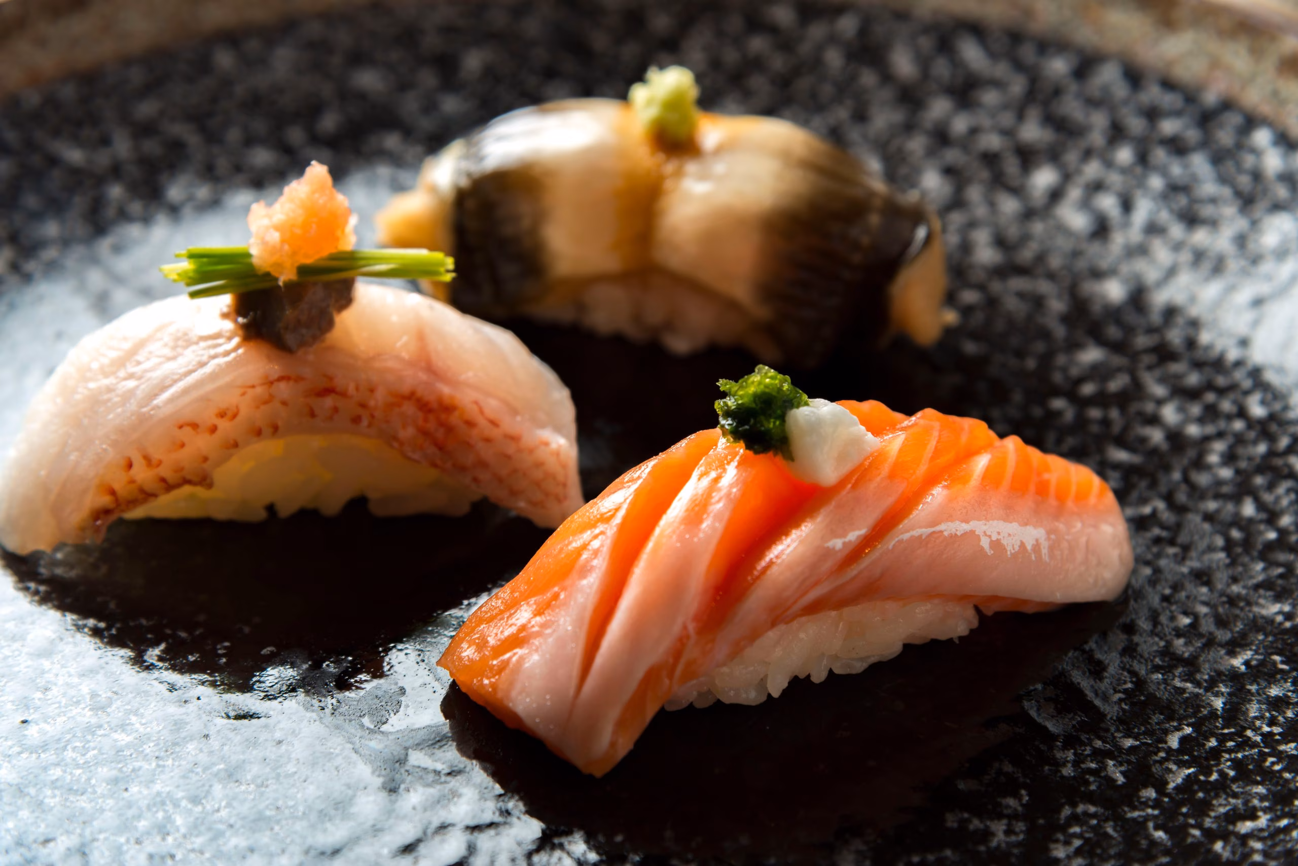 Three pieces of nigiri sushi on a dark, textured plate.