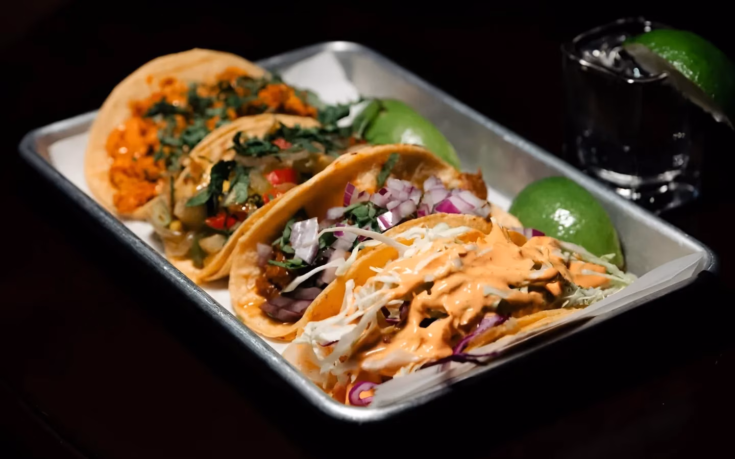 Four tacos, lime wedges, and a shot glass on a metal tray.