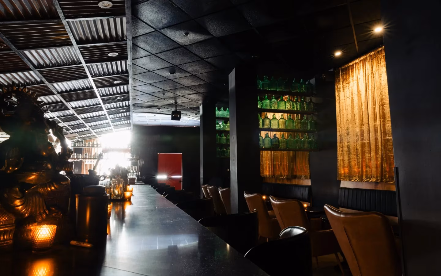 Dark bar interior with counter, green bottles, and leather chairs.
