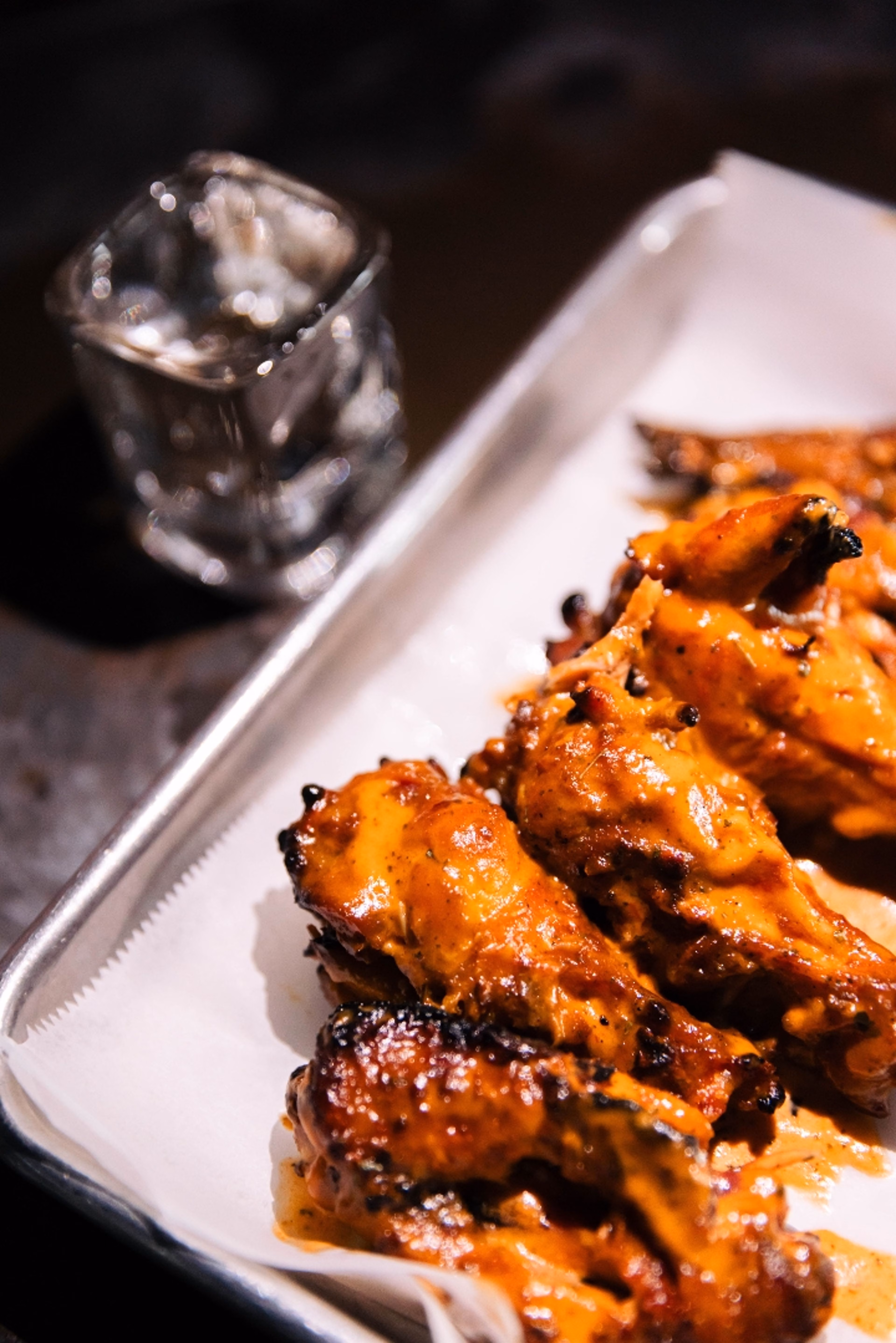 Saucy chicken wings on a metal tray with a blurred glass.