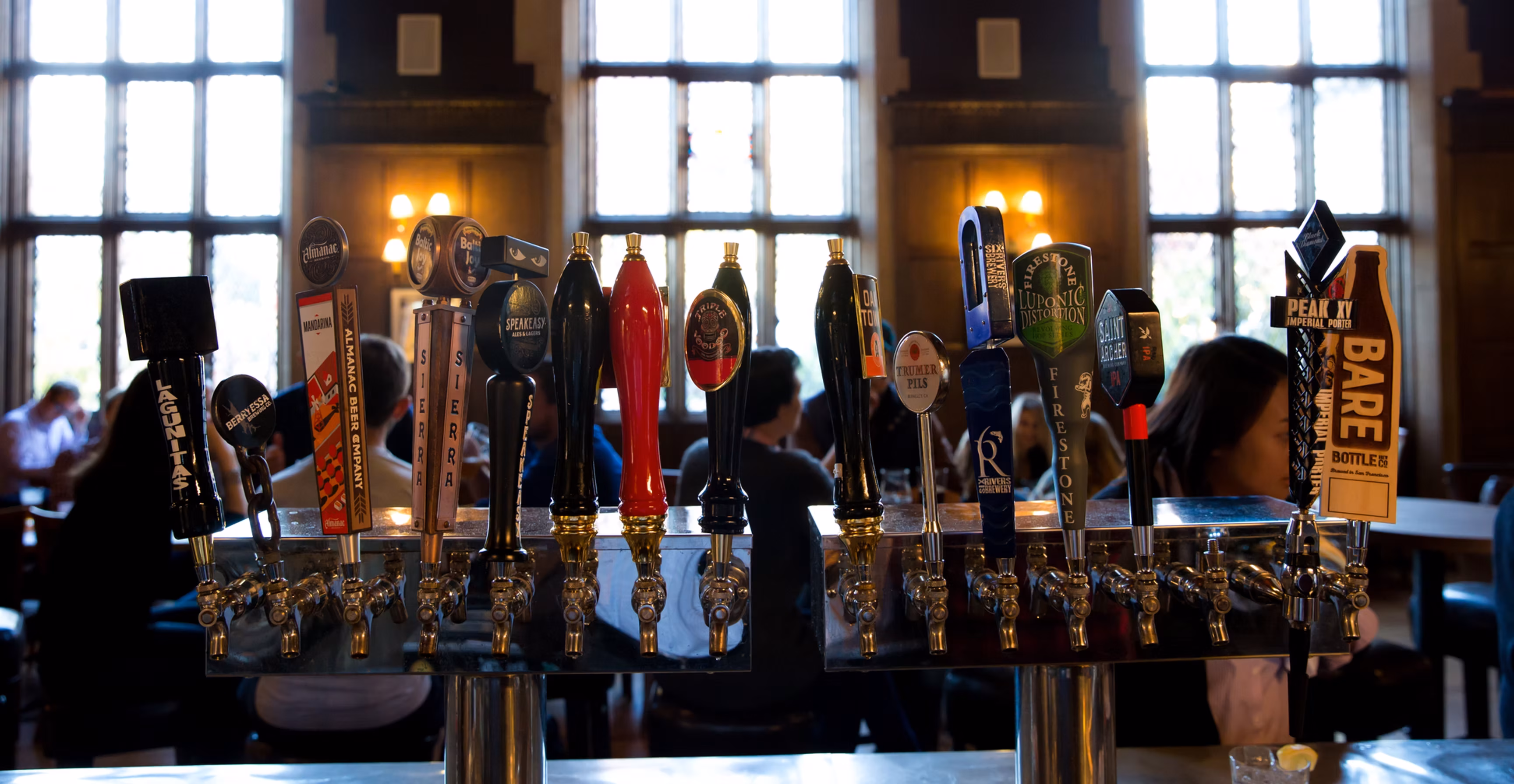 Many beer taps on a bar with large windows in background.