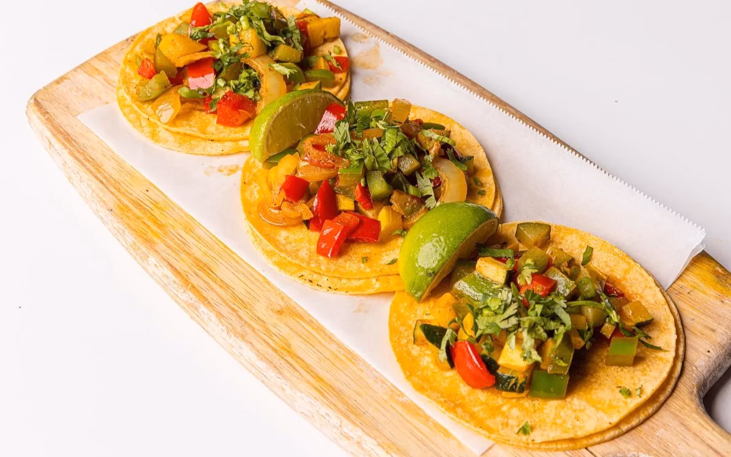 Three vegetable tacos with lime and cilantro on a wooden board.