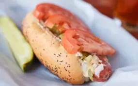 Hot dog with relish and tomatoes, beside a dill pickle spear.
