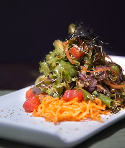 Green salad with red cubes, orange strands, shredded dark topping and sesame seeds.