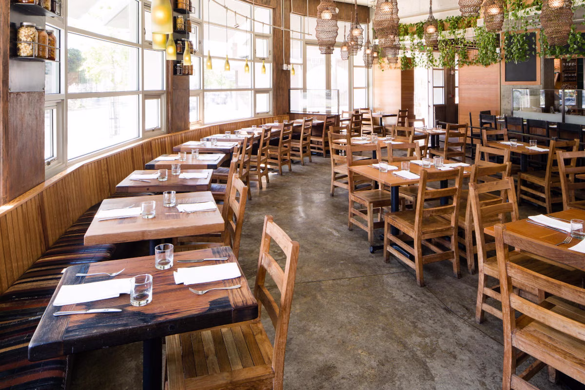 Rustic restaurant interior with wooden tables, chairs, large windows, and plants.