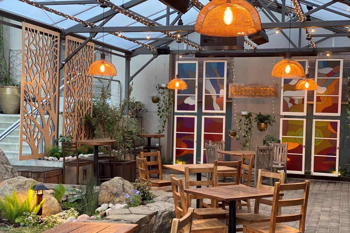 Restaurant patio with wooden tables, tree screens, and wicker lamps.