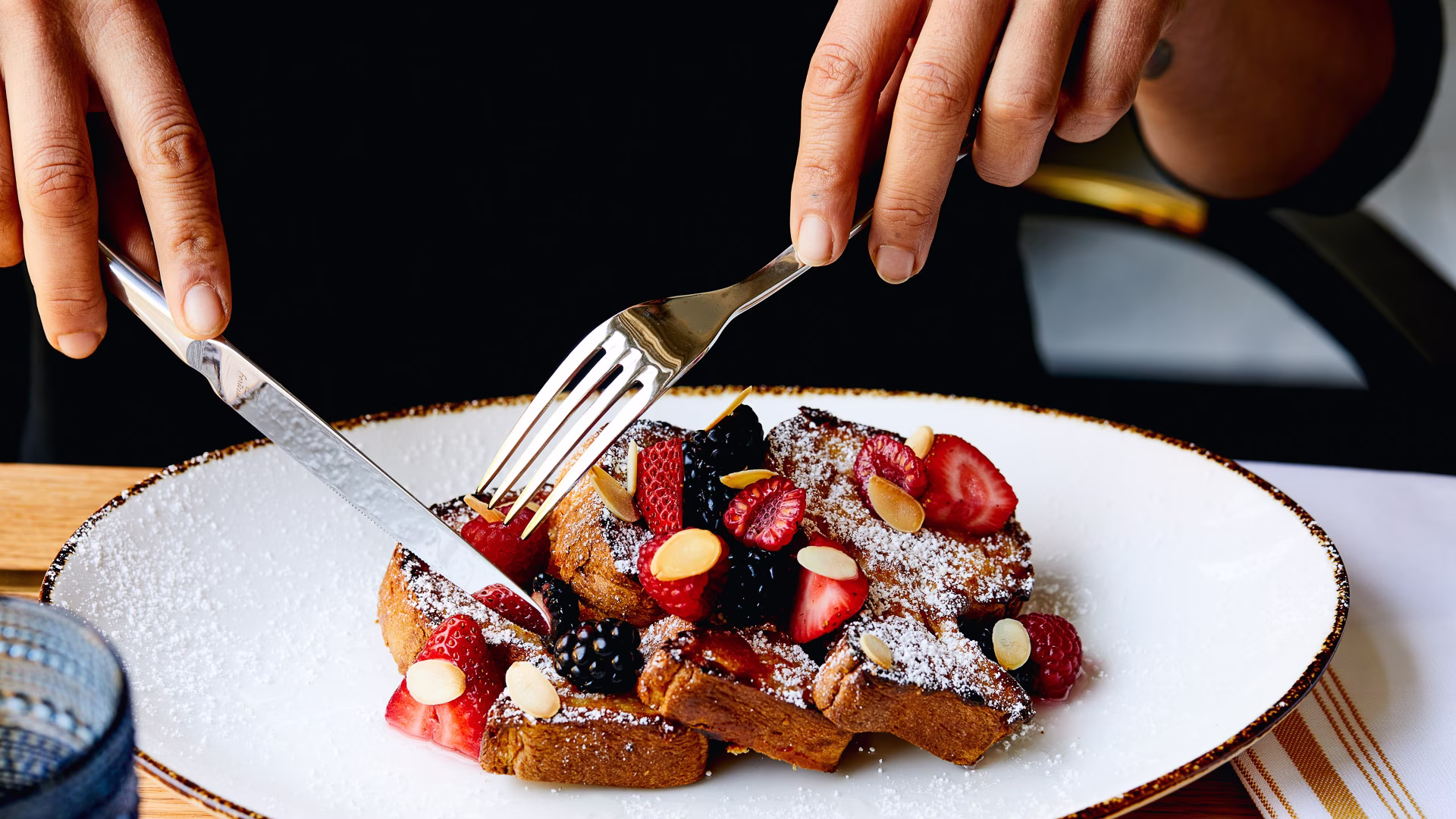 Person cutting French toast with berries, powdered sugar, and almonds.