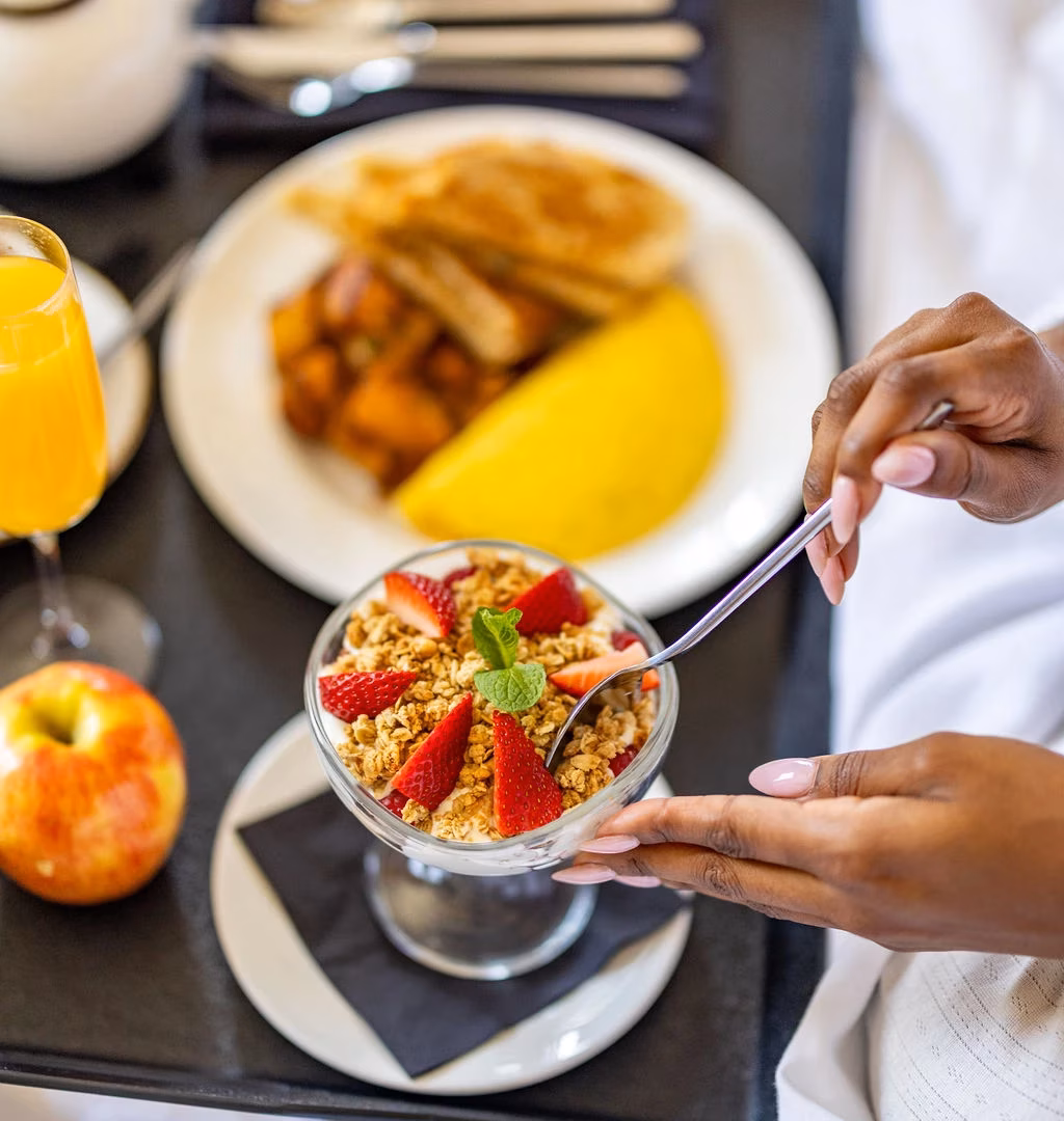 Hands spooning strawberry granola yogurt parfait, with a breakfast spread.