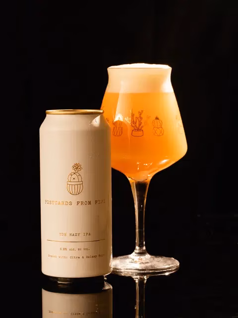 White beer can next to a glass of hazy IPA with cactus art.