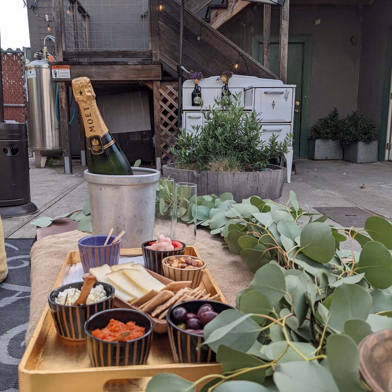 Outdoor table with champagne bottle, snacks, and eucalyptus leaves.