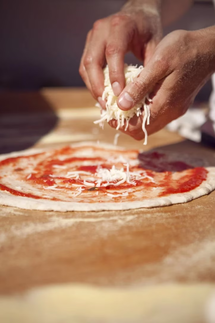 Hands sprinkling shredded cheese onto a pizza base with tomato sauce.