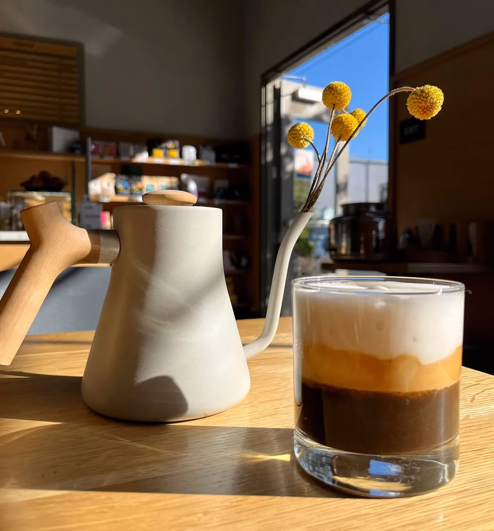 Gooseneck kettle, layered coffee drink, yellow flowers on a sunny wooden table.