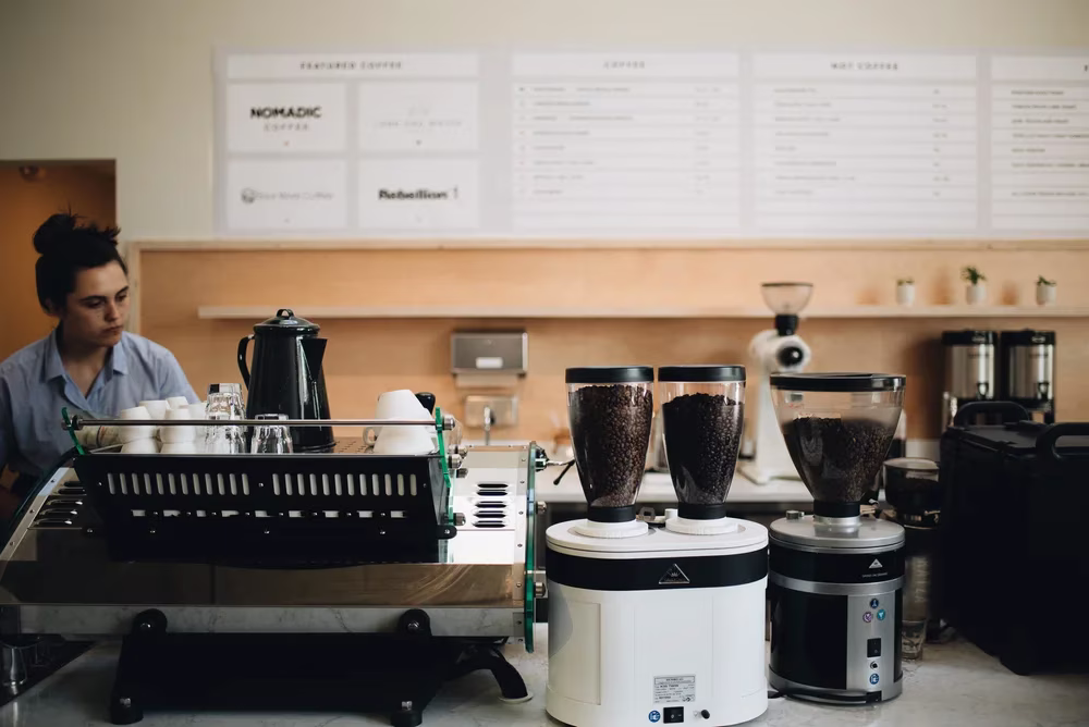 Barista behind counter with coffee grinders and espresso machine.