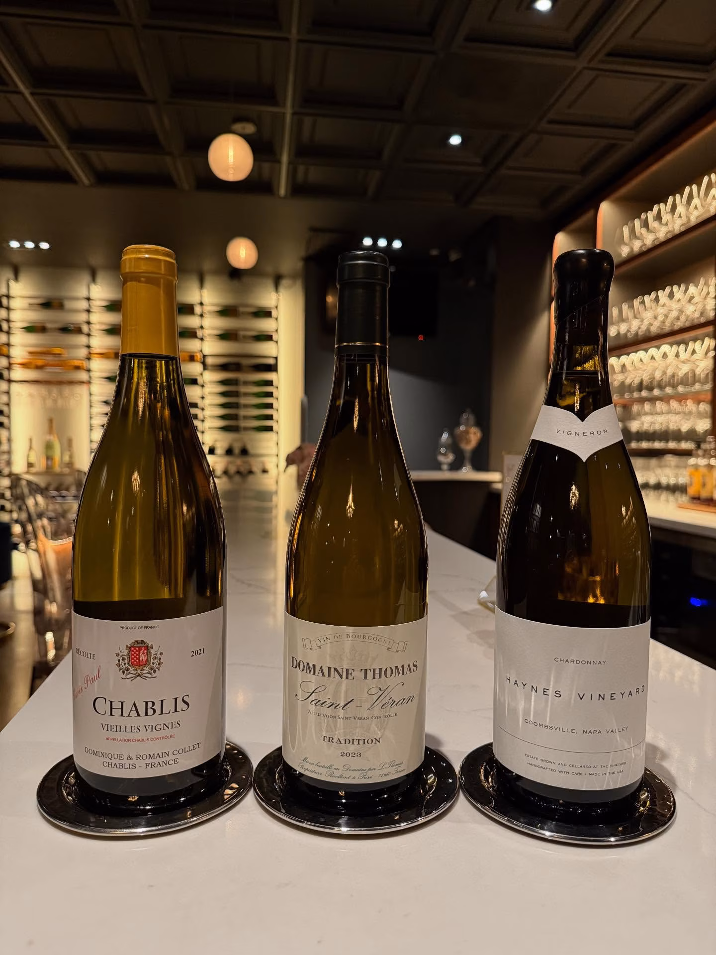 Three bottles of Chablis, Saint-Veran, and Chardonnay on a bar counter.