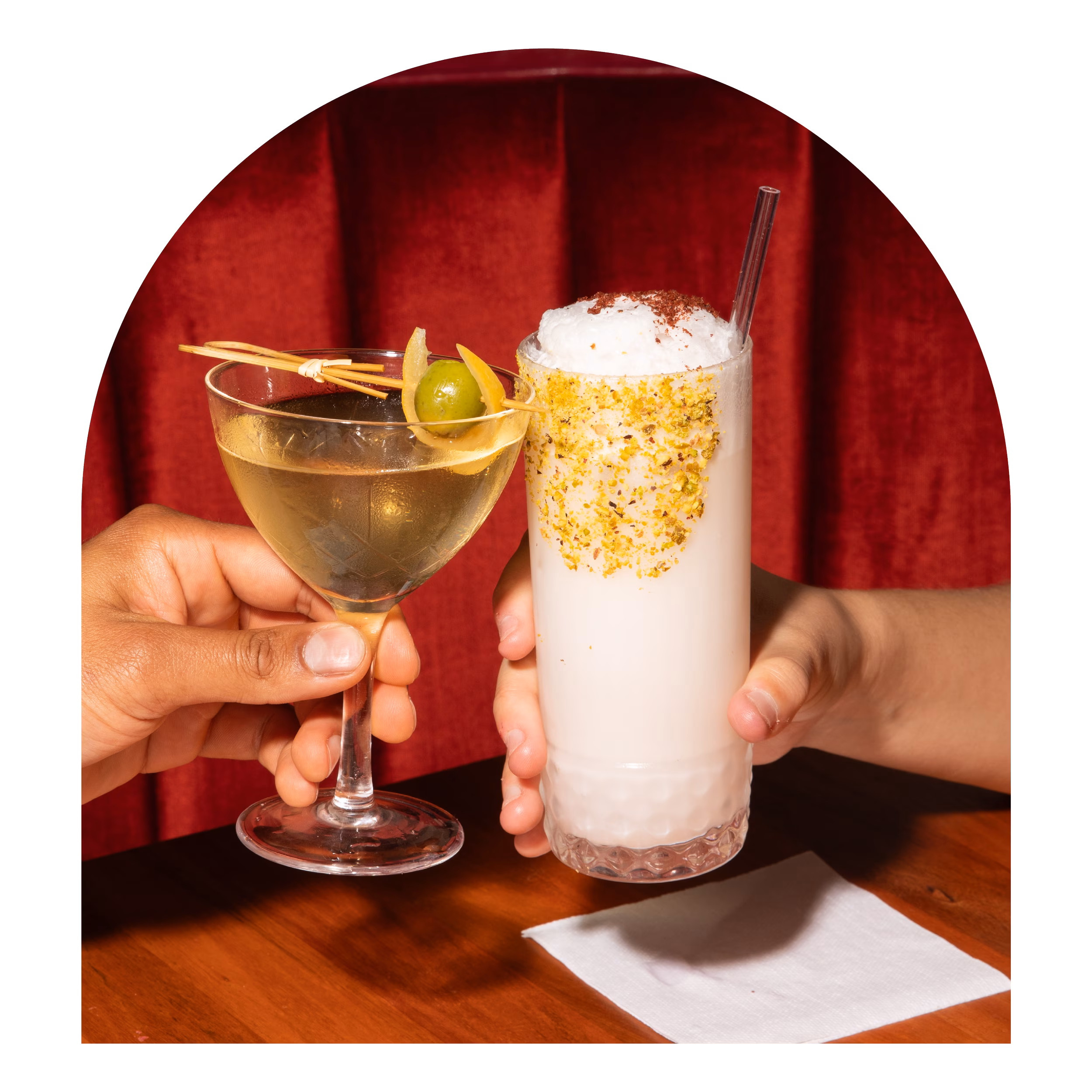 Two hands hold two different cocktails against a red backdrop.