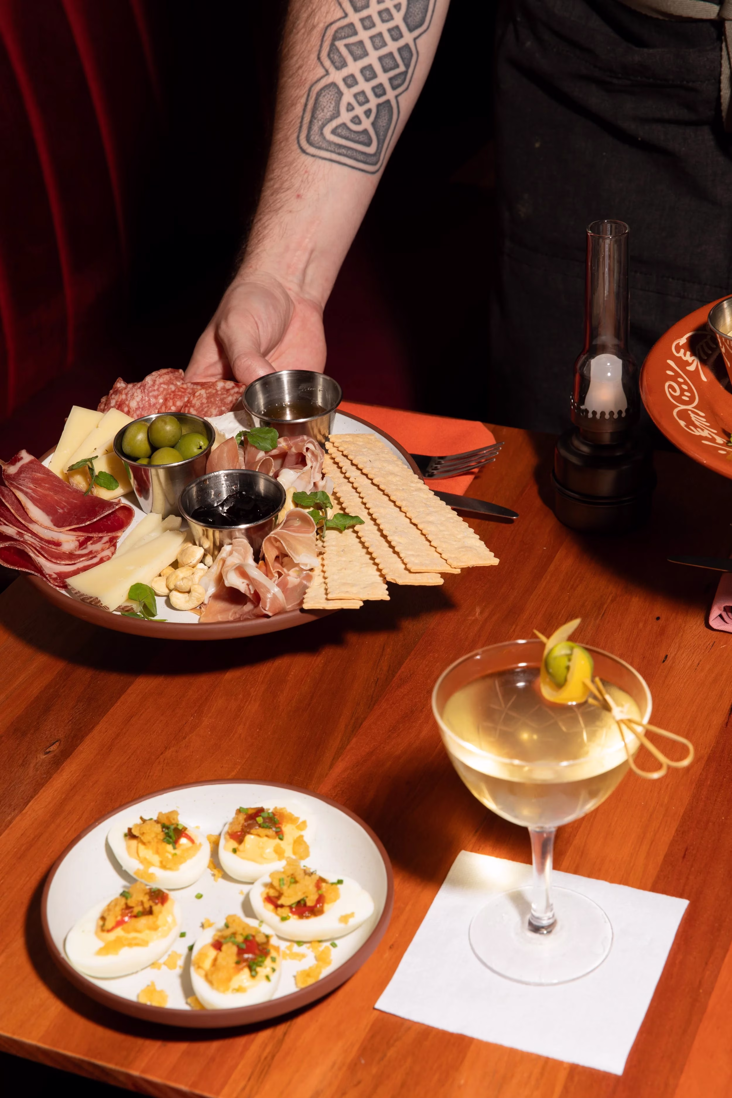 Person serving charcuterie, deviled eggs, and cocktail on a wooden table.