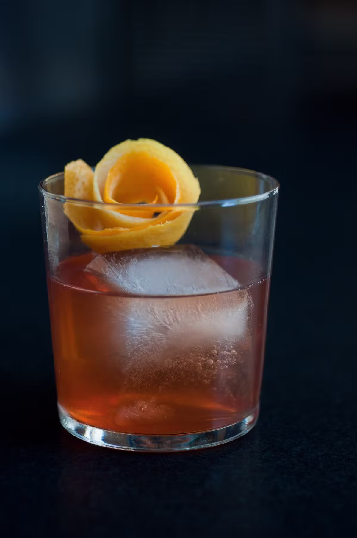 A reddish drink with a large ice cube and orange peel.