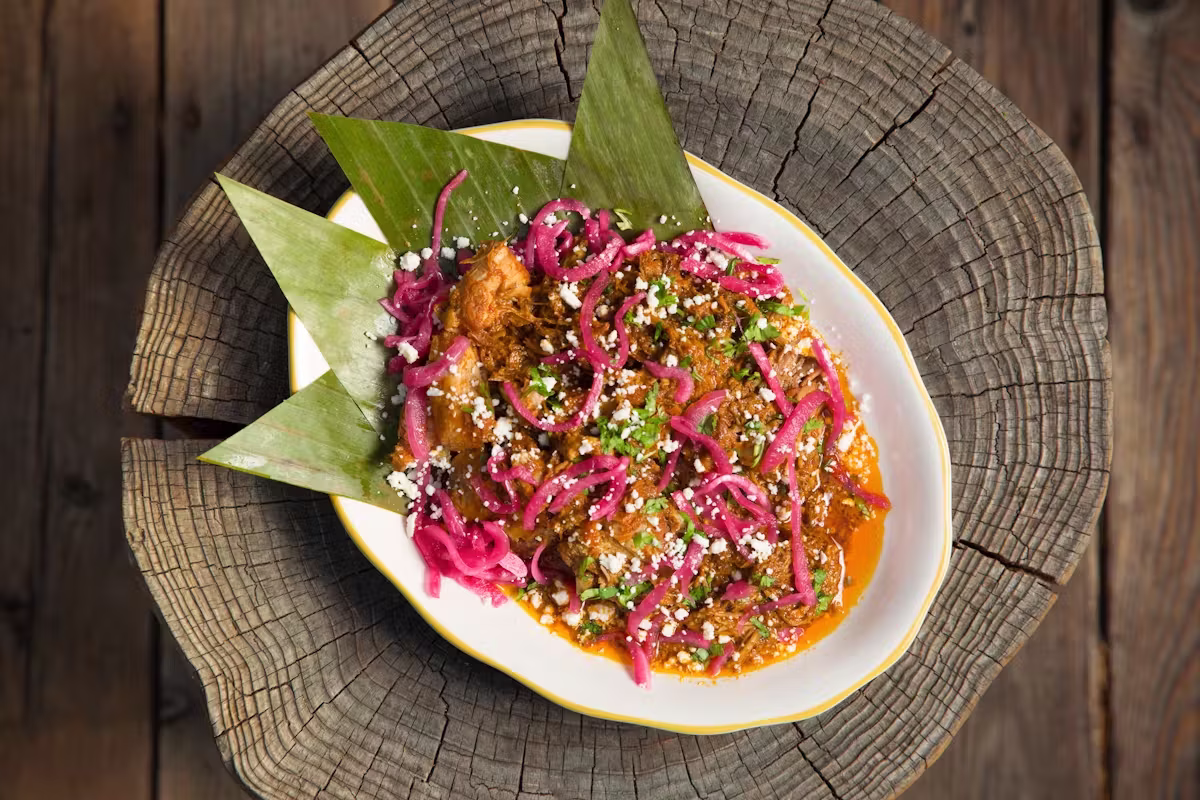 Stewed meat with pickled red onions, cheese, and banana leaves on wood.