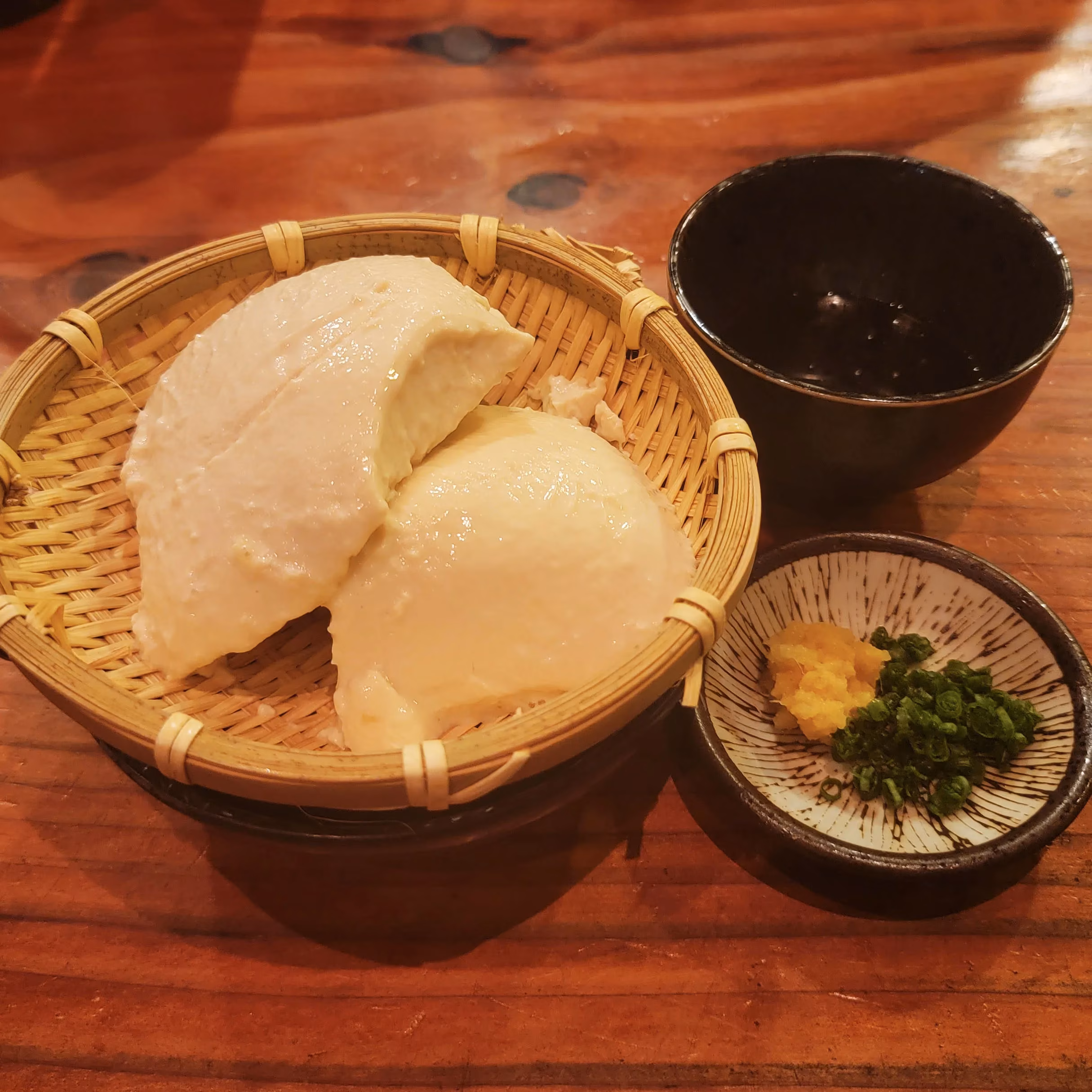 Two pieces of tofu in a basket with dipping sauce and greens.