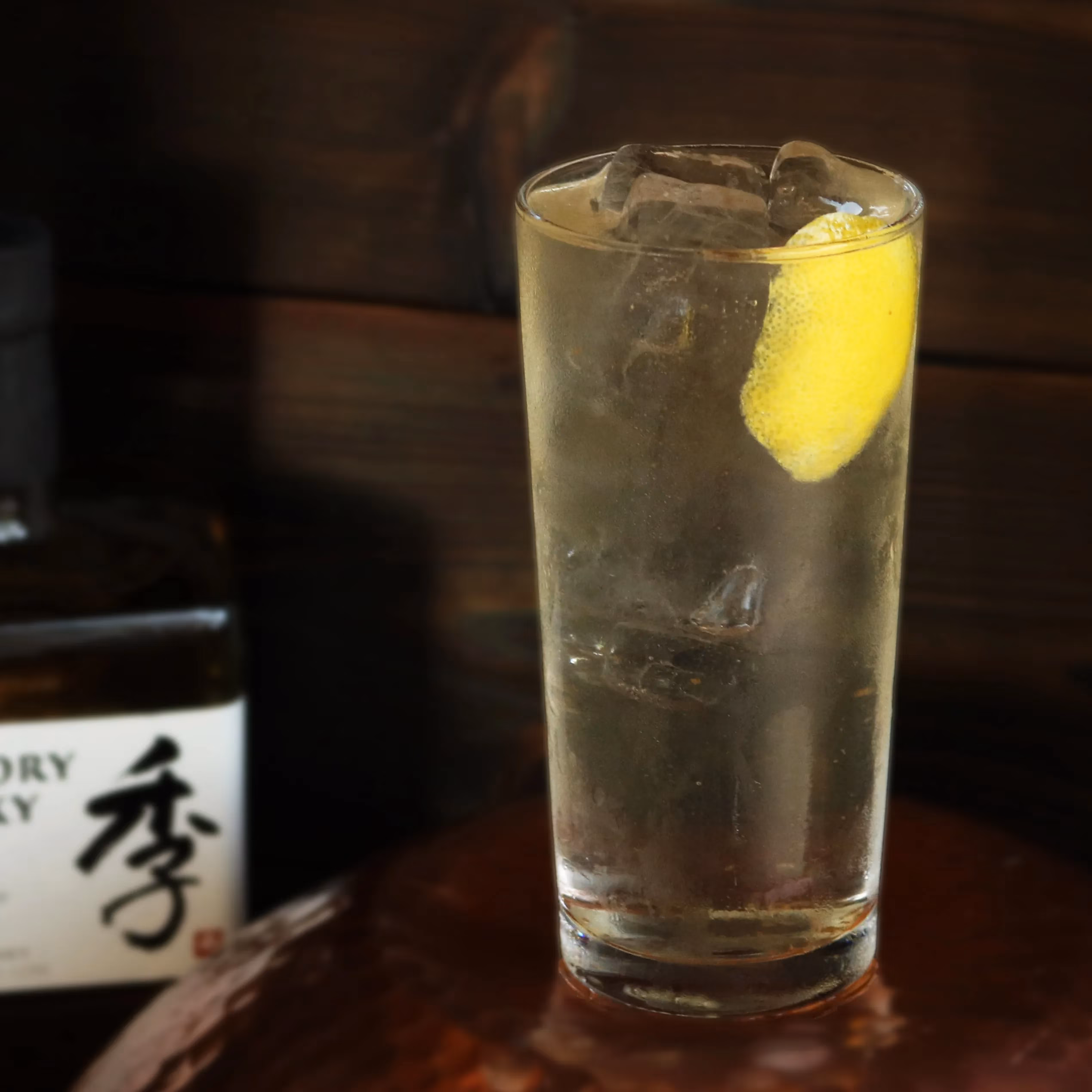 Tall glass of iced clear drink with lemon and blurred bottle.