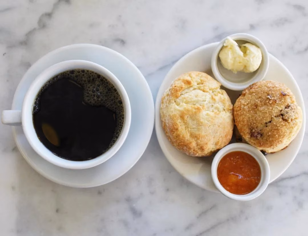 Black coffee, two scones, butter, and jam on a marble table.