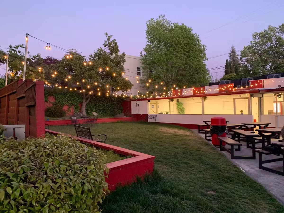 Grassy outdoor patio with string lights, picnic tables, and red trim.