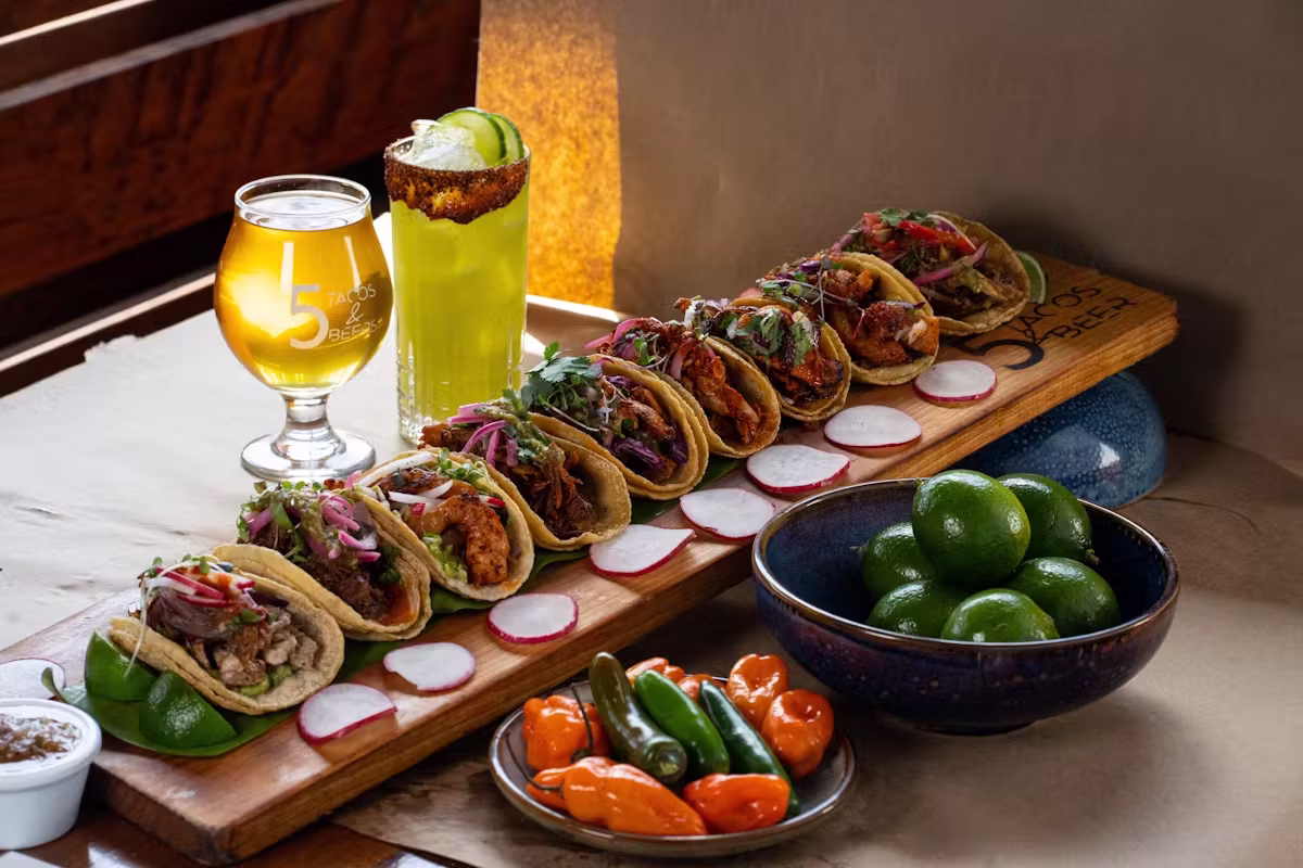 Tacos, beer, cocktail, limes, and radishes on a wooden board.