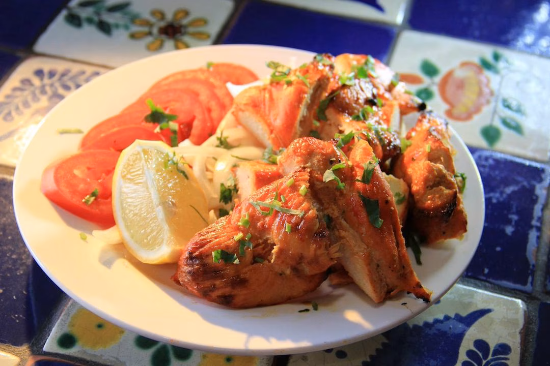 Grilled chicken pieces with tomatoes, lemon, onions, and herbs on a plate.