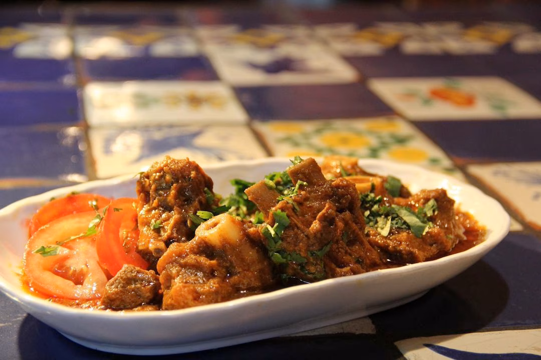 Meat and bone curry with tomatoes and cilantro in a white bowl.
