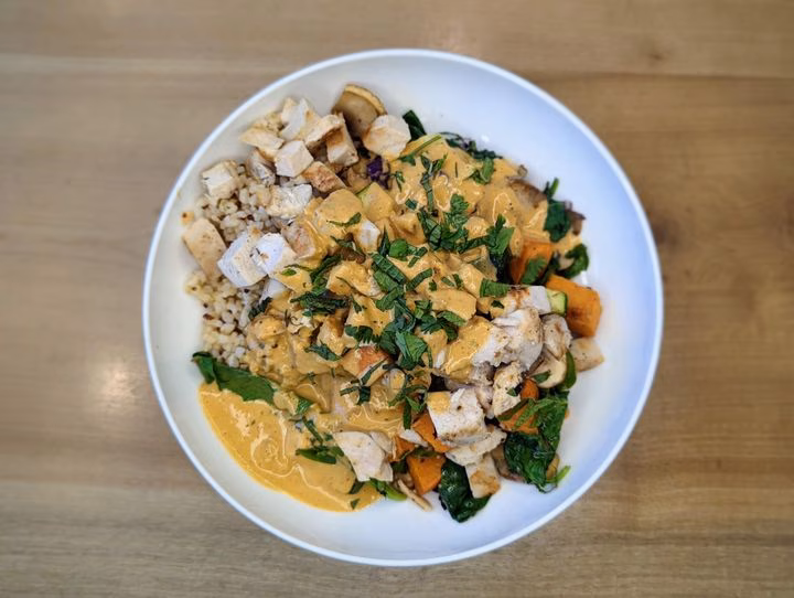 White bowl filled with chicken, grains, vegetables, and creamy sauce.