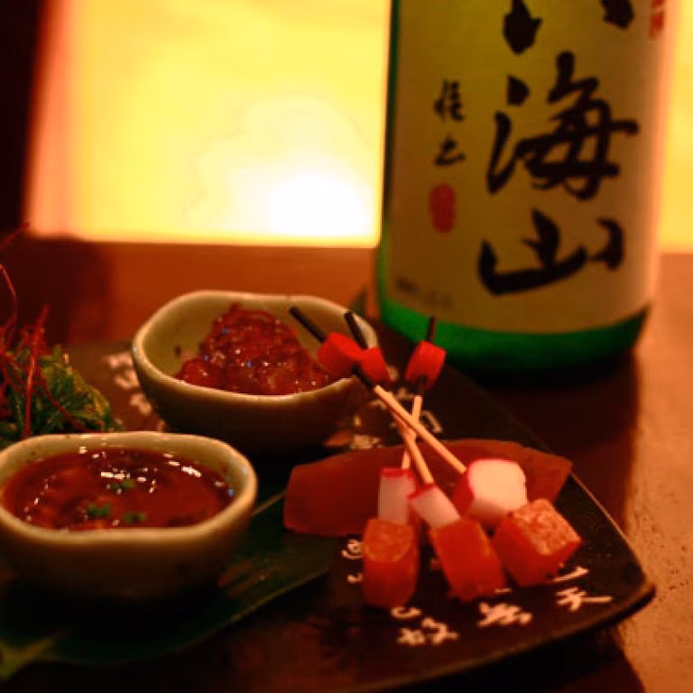 Japanese bottle, two sauces, and skewered appetizers on a dark table.