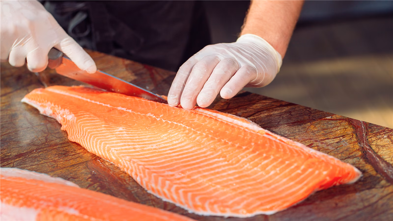 Gloved hands slicing a large orange salmon fillet with a knife.