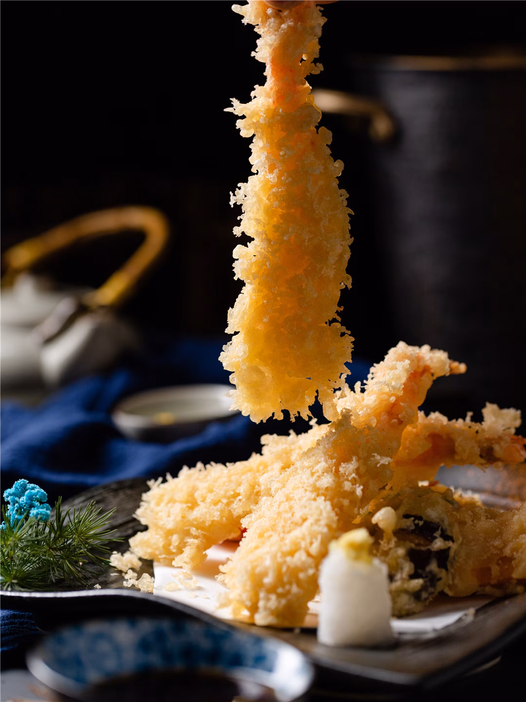 A hand lifts a piece of golden shrimp tempura from a plate.