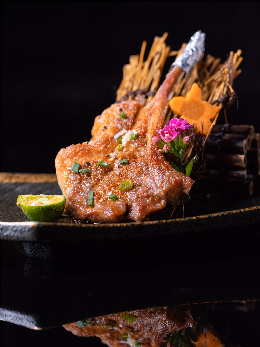 Seasoned lamb chop, lime, and flower elegantly presented on a dark plate.