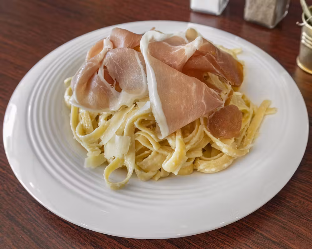 Plate of pasta topped with prosciutto and shaved cheese.
