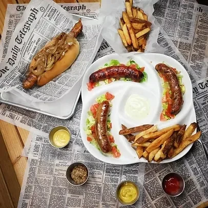 A spread of hot dogs, sausages, fries, and condiments on newspaper placemats.