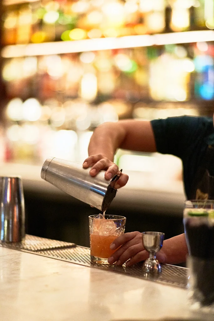 Bartender pours orange drink from shaker into ice-filled glass.