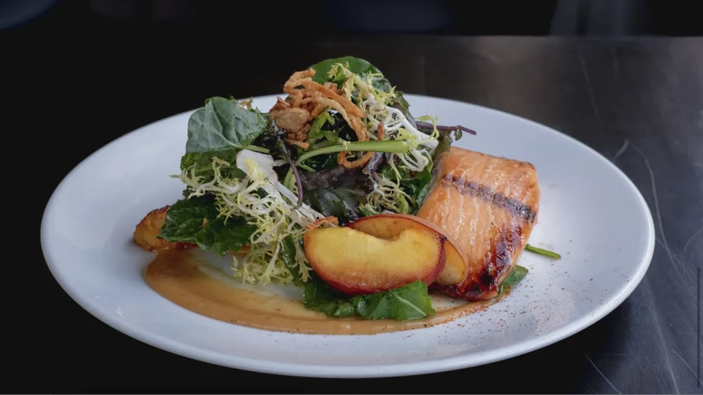 Grilled salmon, mixed greens, grilled peaches, and sauce on white plate.