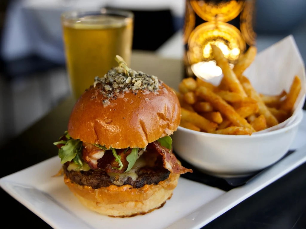 Gourmet burger with bacon, cheese, arugula, and golden fries on a white plate.