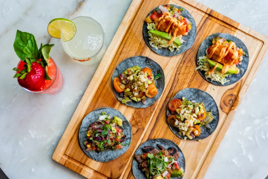 Six blue corn tacos and two garnished cocktails on a wooden board.