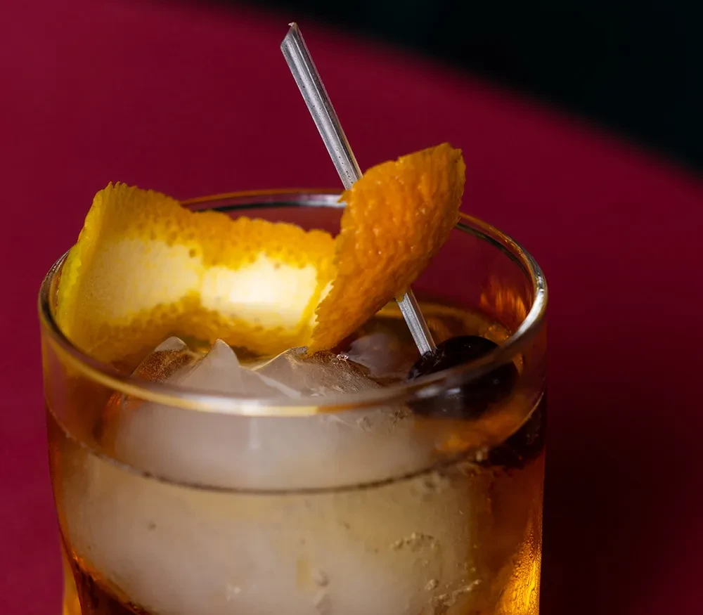 Amber-brown cocktail with ice, orange peel, and cherry in old-fashioned glass.