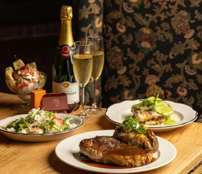 Lavish dining table with steak, champagne, salad, sundae, and gift.