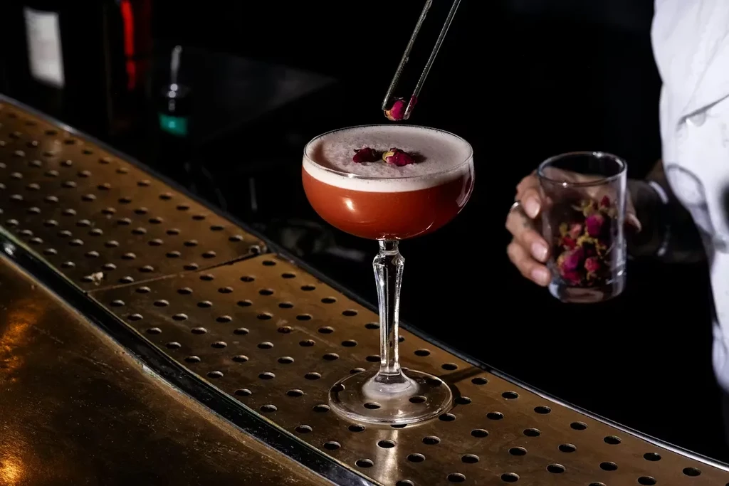 A coupe cocktail with foam being garnished with a dried rose petal.