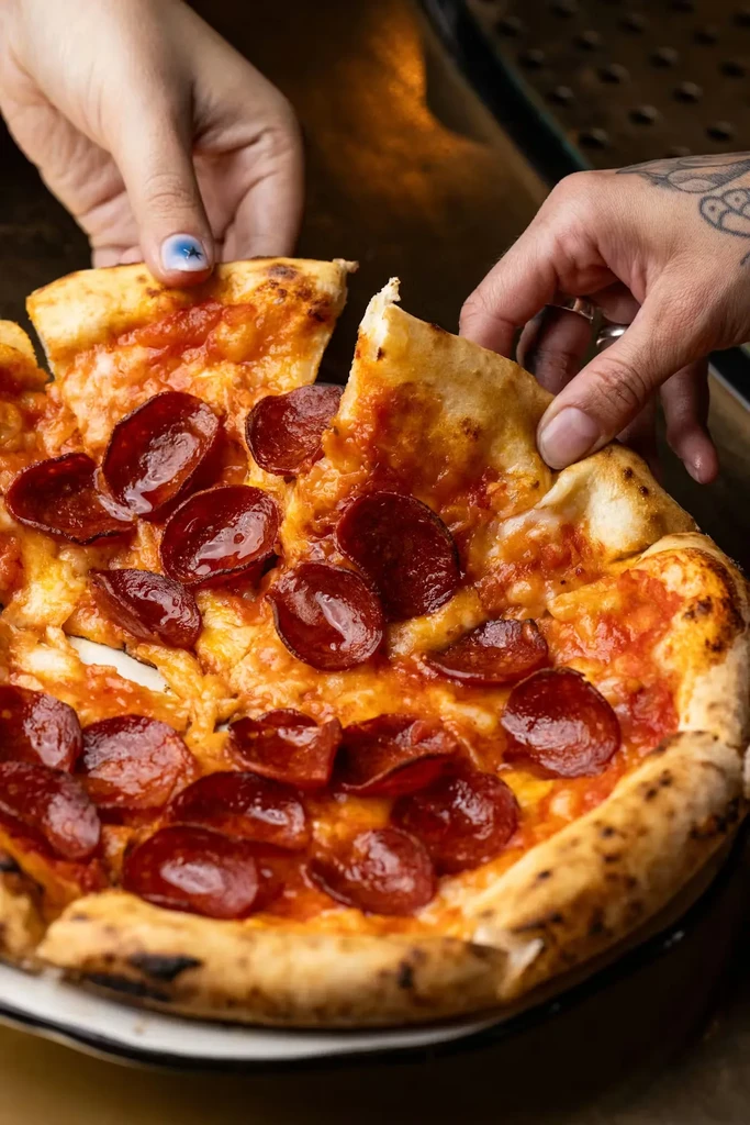 Two hands pulling cheesy pepperoni pizza slices apart.